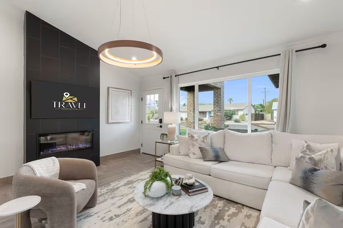 Sunlit modern suburban living room with a white L-shaped sectional, round marble coffee table, circular pendant light, wall fireplace and large front window overlooking the neighborhood.