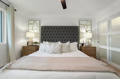 Bright, cozy modern master bedroom with a tall tufted charcoal headboard, white linens and beige throw, matching wooden nightstands with lamps, decorative mirrors, frosted sliding glass door and a large curtained window.