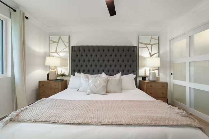 Bright, cozy modern master bedroom with a tall tufted charcoal headboard, white linens and beige throw, matching wooden nightstands with lamps, decorative mirrors, frosted sliding glass door and a large curtained window.