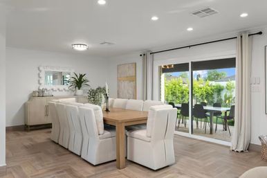 Bright, airy modern dining room with a wooden table surrounded by plush white upholstered chairs, sliding glass doors opening to a sunny backyard patio with a black outdoor dining set and lush green hedges