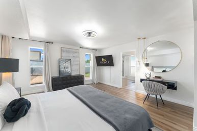 Sunlit contemporary master bedroom with hardwood floors, white bedding and gray throw, knot accent pillow, wall-mounted TV, black dresser with framed art, two tall windows with curtains, round mirror above a floating desk and an en-suite bathroom visible