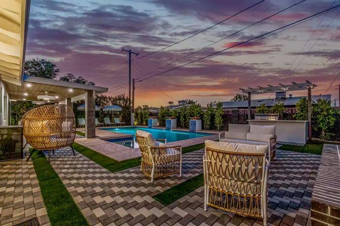 Modern residential backyard pool patio at sunset with a rectangular pool, wicker lounge chairs and hanging egg chair, pergola and outdoor bar on patterned pavers.