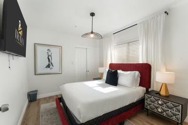 Bright, modern bedroom with red upholstered headboard, crisp white bedding, black accent pillow, gold-accent lamps, patterned nightstand, framed art, wall-mounted TV and sunlit window with white curtains.