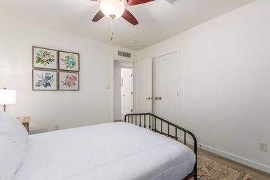 Bright, minimalist bedroom interior with white walls and bedding, black metal bed frame, four-piece floral wall art, wooden nightstand with lamp, ceiling fan with wooden blades, and neutral area rug.