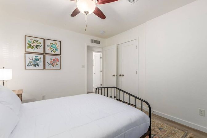 Bright, minimalist bedroom interior with white walls and bedding, black metal bed frame, four-piece floral wall art, wooden nightstand with lamp, ceiling fan with wooden blades, and neutral area rug.