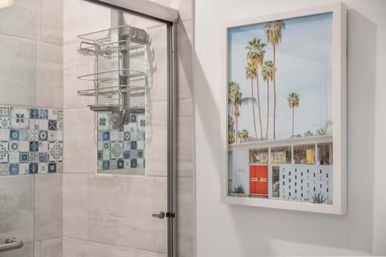 Modern bathroom with glass shower door, blue patterned tile accent and chrome shower caddy, styled with a framed Palm Springs–style photo of tall palm trees and a red front door.