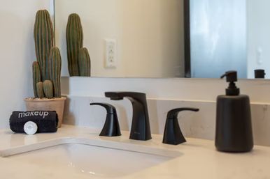 Modern bathroom vanity with white countertop, matte-black three-piece faucet and soap dispenser, potted cactus and rolled 'makeup' towel in front of mirror.