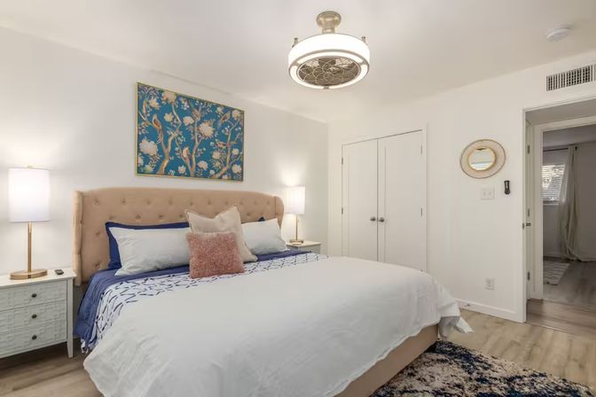 Bright modern master bedroom interior with tufted beige headboard, king bed dressed in white and navy linens, floral wall art, bedside lamps, ceiling light and light wood floors