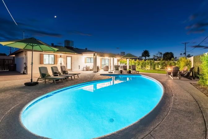 Glowing kidney-shaped backyard swimming pool at dusk beside a single-story home, with lounge chairs and umbrellas, patio seating and a fire pit, illuminated landscaping and a crescent moon overhead.