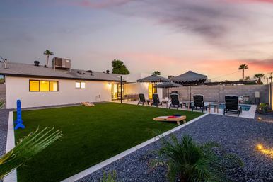 Sunset suburban backyard with artificial turf lawn and cornhole boards, gravel landscaping, poolside lounge chairs under scalloped umbrellas, small swimming pool, single-story stucco house and palm trees — ready for poolside fun.