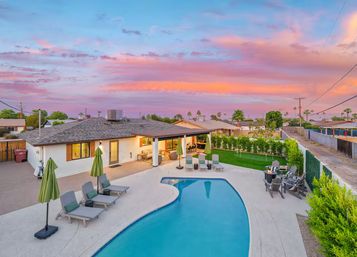 Vibrant pink-orange sunset over a suburban backyard with a kidney-shaped pool, lounge chairs, covered patio, green lawn and palm trees.