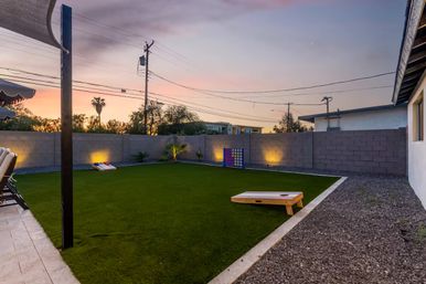 Twilight suburban backyard with artificial turf, cornhole boards and oversized Connect Four game, concrete block wall, palm tree silhouette and warm accent lighting
