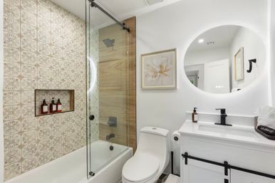 Bright modern bathroom interior with patterned tile accent wall in tub-shower, frameless sliding glass door, wood-look shower panel, white tub and toilet, backlit round mirror and white vanity with matte black fixtures.