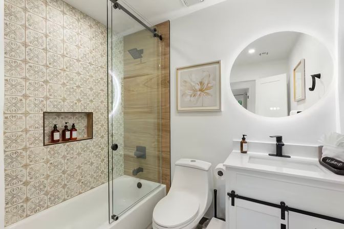 Bright modern bathroom interior with patterned tile accent wall in tub-shower, frameless sliding glass door, wood-look shower panel, white tub and toilet, backlit round mirror and white vanity with matte black fixtures.