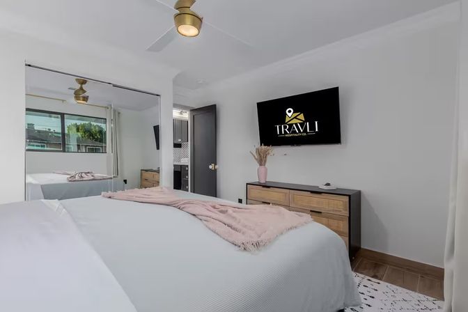 Modern city apartment bedroom — king bed with pale bedding and pink throw, mirrored closet, wooden dresser with wall-mounted TV, brass ceiling fan and urban window view.