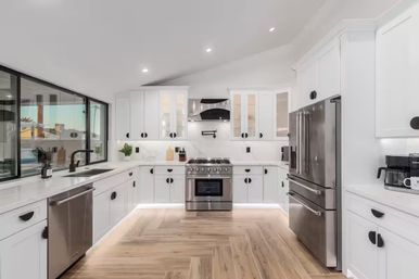 Bright modern U-shaped white kitchen with stainless steel appliances, large window, wood-look floors, and LED under-cabinet lighting.