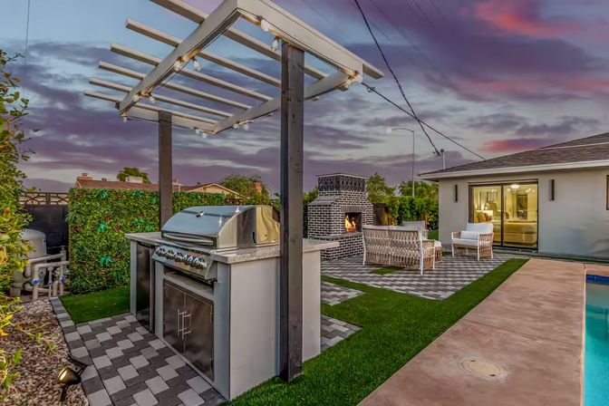 Modern twilight backyard patio with stainless steel built-in grill under a pergola strung with lights, brick outdoor fireplace, wicker seating, pool, and sliding glass doors