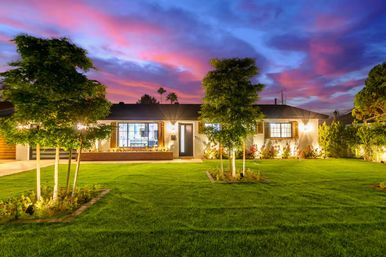 Twilight curb appeal of a single-story suburban ranch home with warm exterior lights, a manicured green lawn, young ornamental trees and landscaped flower beds beneath a pink-purple sunset sky.