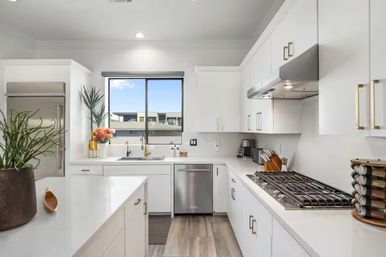 Bright, sunlit modern white kitchen with stainless steel refrigerator, dishwasher and gas cooktop, large island, potted plants and a window overlooking nearby rooftops and blue sky