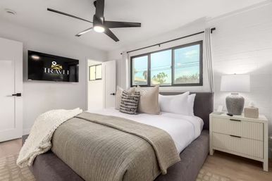 Sunlit modern vacation rental bedroom with king bed, neutral linens, textured throw and pillows, wall-mounted TV, bedside lamp, and ceiling fan