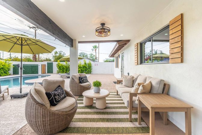 Covered backyard patio with round wicker chairs, beige sofa on a striped rug, coffee table, green umbrellas and an adjacent swimming pool
