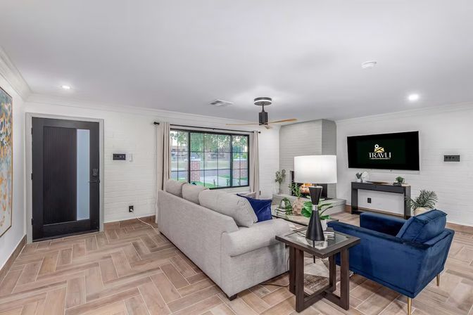 Bright modern living room with light-gray sofa, navy velvet armchairs, wood-look herringbone tile floor, large front window, fireplace, ceiling fan and black entry door.