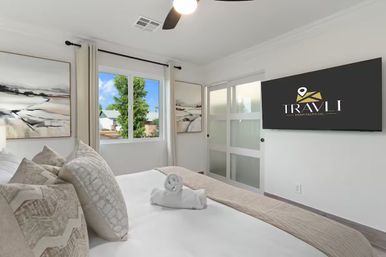 Sunlit modern bedroom in neutral tones with plush pillows and a rolled towel on a neatly made bed, abstract wall art, wall-mounted flat-screen TV, frosted sliding door, ceiling fan and a window view of a tree.