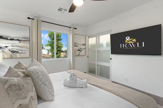 Sunlit modern bedroom in neutral tones with plush pillows and a rolled towel on a neatly made bed, abstract wall art, wall-mounted flat-screen TV, frosted sliding door, ceiling fan and a window view of a tree.