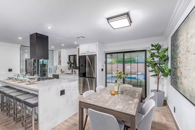 Bright modern open-concept kitchen and dining area with white marble island and gray bar stools, stainless steel refrigerator and black range hood, wooden dining table with chairs, sliding glass doors to a backyard pool, large abstract wall art and potted plant.