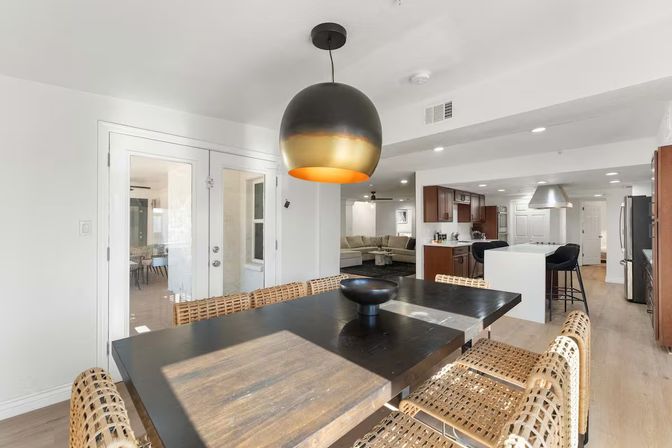 Sunlit open-plan dining area with dark wood table, woven rattan chairs, large black-and-gold pendant light, and view into a modern kitchen with island and living room