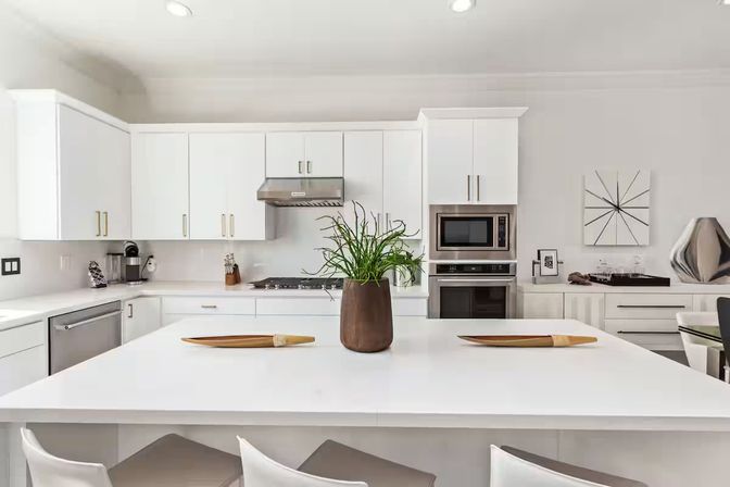 Bright modern white kitchen with large island, stainless steel range, built-in microwave, wooden vase with green plant and bar stools