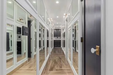 Long luxury walk-in closet with floor-to-ceiling mirrored wardrobe panels, warm wood-look tile floor, recessed ceiling lights, brass hardware and dark built-in cabinetry at the far end.