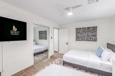 Bright modern short-term rental bedroom with two neatly made beds, white linens and blue accent pillows, ceiling fan, wall-mounted TV, mirrored closet doors, and abstract wall art.