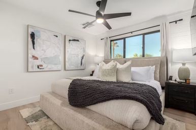 Bright modern bedroom with upholstered king bed, neutral pillows and chunky dark-gray throw, abstract wall art, black ceiling fan, bedside lamps, and large window with sunny palm-tree view — coastal, contemporary style.