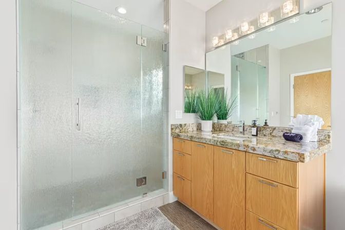 Bright contemporary bathroom with textured glass walk-in shower, granite countertop and wood vanity, large mirror with vanity lights, potted plant and folded towels