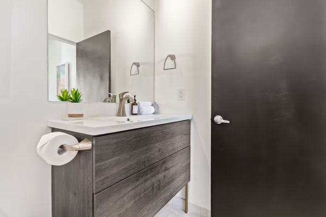 Sleek modern bathroom vanity with dark wood drawers, white countertop sink, brushed-nickel faucet and toilet-paper holder, mirror, rolled towel, small succulent, and matte black door.