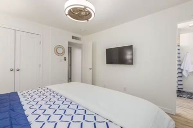 Bright modern bedroom with white walls, wall-mounted flat-screen TV, blue geometric bedding on a neatly made bed, round ceiling fan light, and doorway leading to an en-suite bathroom.