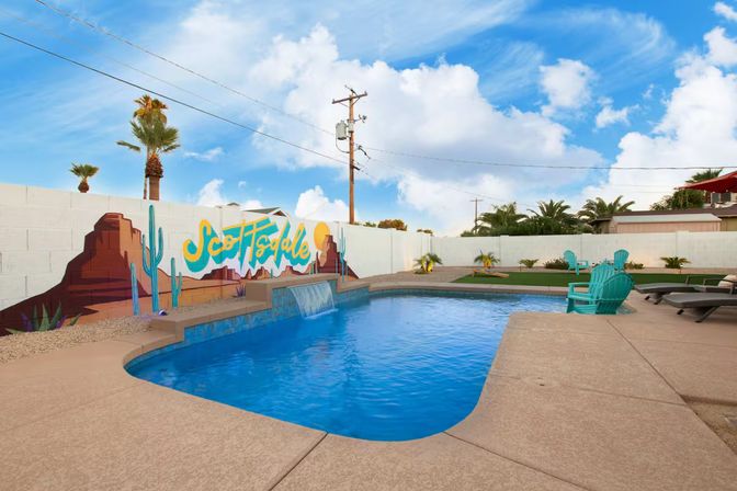 Sunny Scottsdale backyard pool with a small waterfall spillover, turquoise lounge chairs, painted desert mural on a white block wall, palm trees and bright blue sky