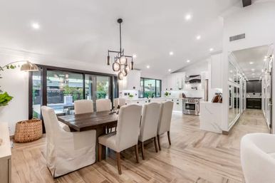 Bright modern open-concept dining room and kitchen with a large wooden table and upholstered chairs, pendant chandelier, white cabinetry, stainless steel range, sliding glass doors to a patio and pool, and light wood floors.