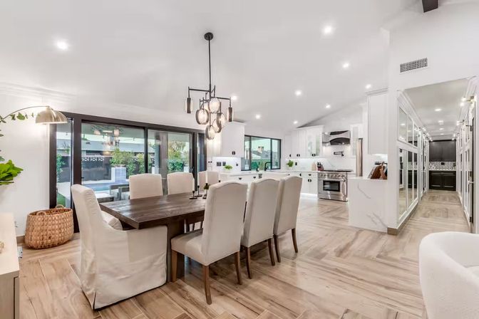 Bright modern open-concept dining room and kitchen with a large wooden table and upholstered chairs, pendant chandelier, white cabinetry, stainless steel range, sliding glass doors to a patio and pool, and light wood floors.