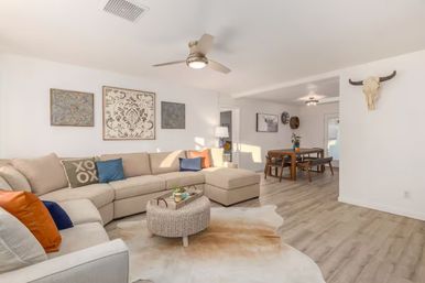 Sunlit open-concept living room and dining area with a beige sectional sofa, colorful throw pillows, woven ottoman on a cowhide rug, light wood floors, wall art, mounted longhorn skull and modern ceiling fan.