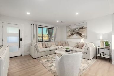 Bright contemporary living room with light-gray L-shaped sectional, round marble coffee table, textured accent chair and neutral rug under large abstract wall art, black-framed window, white entry door and side table with lamp and potted plant — cozy neutral palette and abundant natural light.