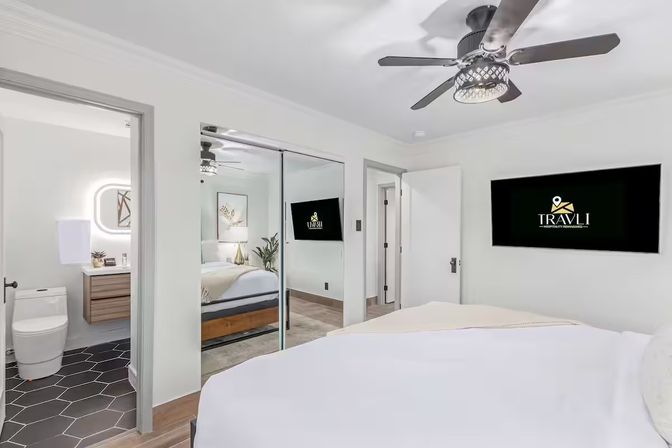 Bright modern hotel bedroom with white walls, ceiling fan, queen bed, mirrored sliding closet, wall-mounted TV, potted plant, and an en-suite bathroom featuring hexagon tile floor and illuminated vanity mirror.