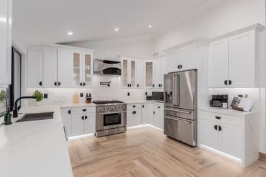 Bright modern white kitchen interior with stainless steel French-door refrigerator and gas range, white shaker cabinets with black pulls, marble-look quartz counters, under-cabinet and toe-kick LED lighting, black faucet and wood-grain tile floor.