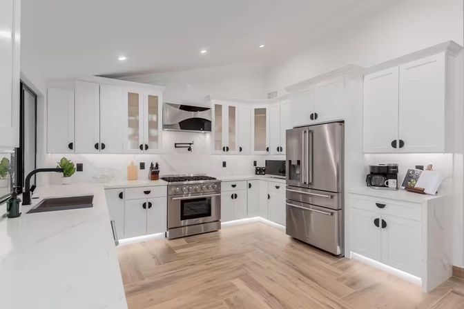 Bright modern white kitchen interior with stainless steel French-door refrigerator and gas range, white shaker cabinets with black pulls, marble-look quartz counters, under-cabinet and toe-kick LED lighting, black faucet and wood-grain tile floor.