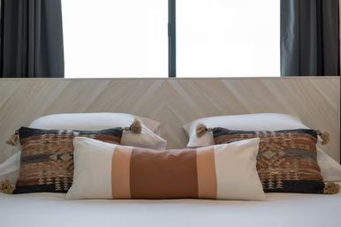 Cozy modern bedroom scene with a chevron wood headboard, central tan-and-cream lumbar pillow flanked by two patterned boho tassel throw pillows, white bedding and dark curtains framing a bright window.