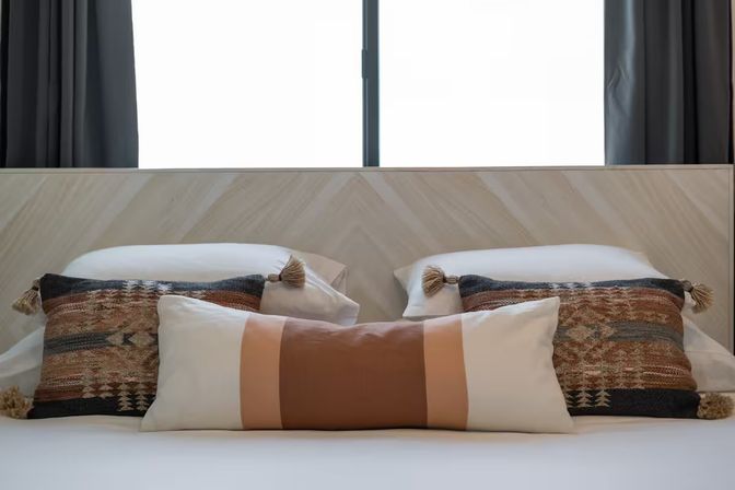 Cozy modern bedroom scene with a chevron wood headboard, central tan-and-cream lumbar pillow flanked by two patterned boho tassel throw pillows, white bedding and dark curtains framing a bright window.