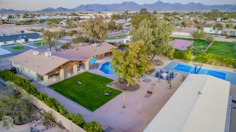 Aerial view of a suburban desert backyard with a covered patio, blue swimming pool, rectangular artificial turf lawn, large shade trees, and a bright blue outdoor basketball court with mountains on the horizon.