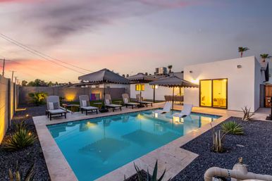 Sunset-lit modern backyard with rectangular swimming pool, in-pool loungers, multiple chaise lounges and scalloped umbrellas, sliding glass door to a white single-story house, and low-maintenance desert landscaping with gravel and agave plants.