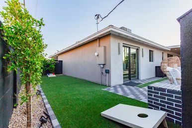 Modern backyard patio of a single-story home with artificial turf, paver pathway, sliding glass doors, wicker egg chair, brick planter and cornhole boards under a clear sky